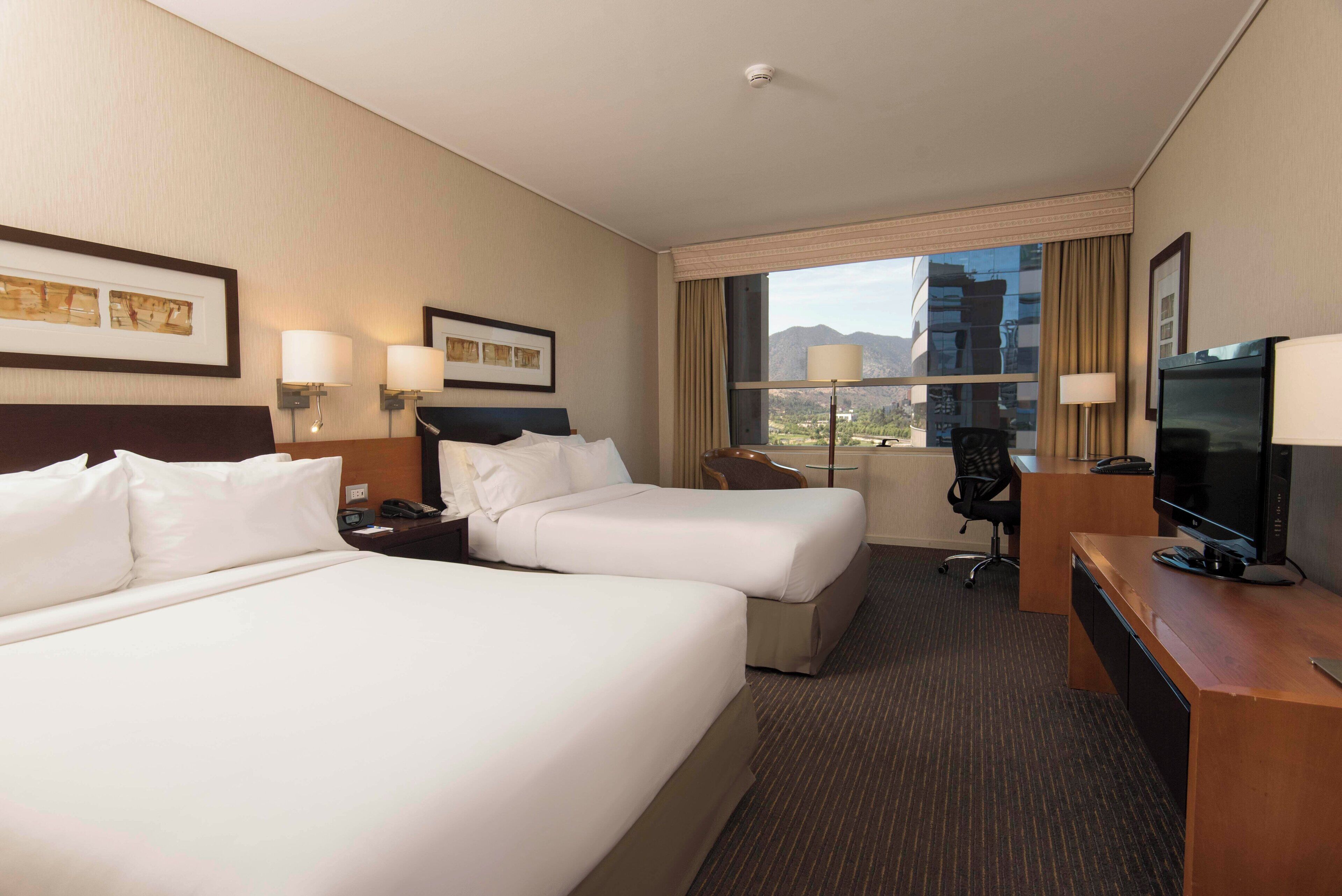 A hotel room with two beds, a desk, a chair, a TV, and a window with a mountain view.
