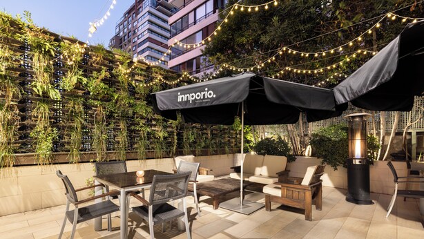 Ein Terrassenbereich im Freien mit Tisch, Stühlen, Lichterketten und einem schwarzen Sonnenschirm mit dem Aufdruck "innporio".