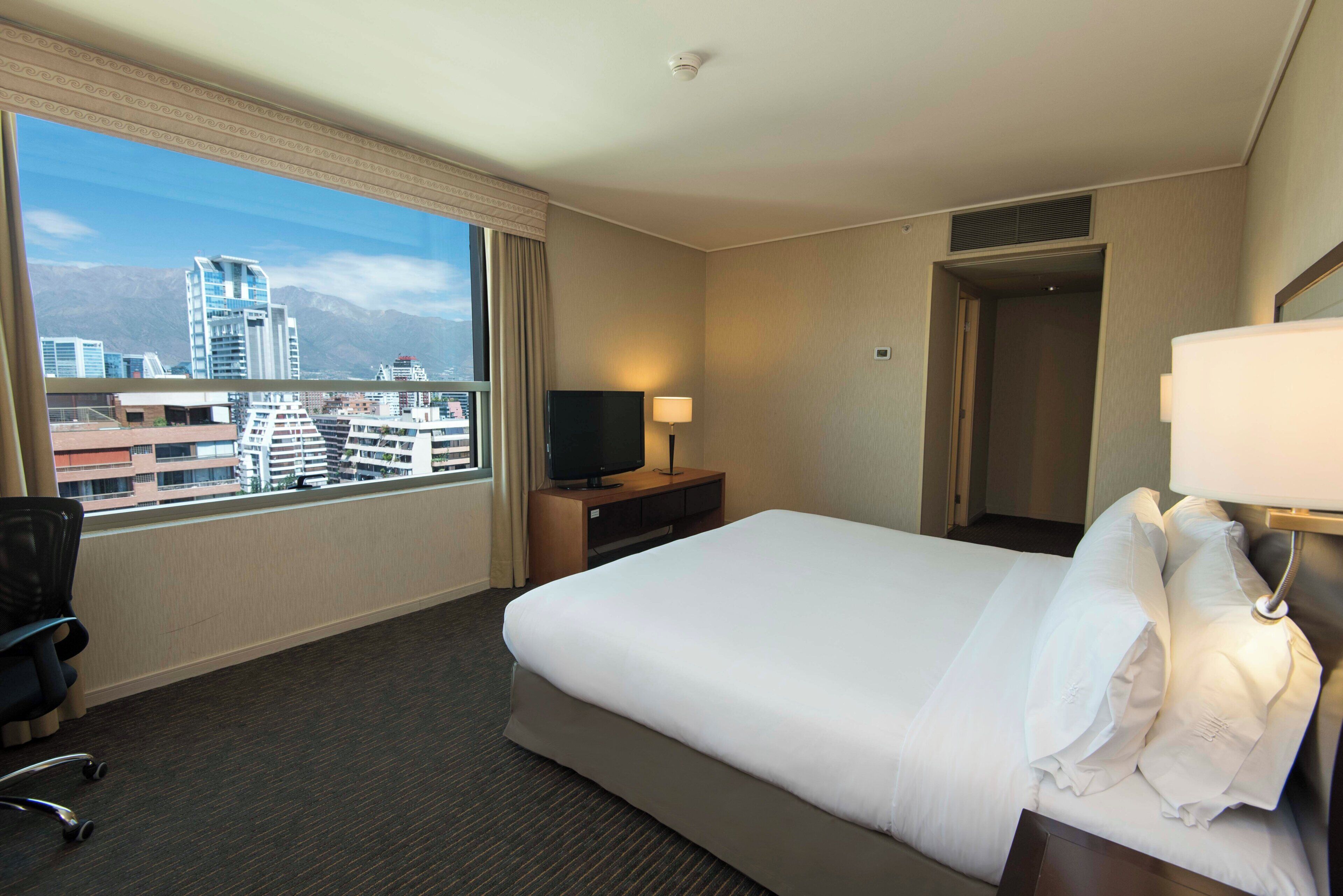 A hotel room with a large bed, a TV, a desk, and a window with a city view.