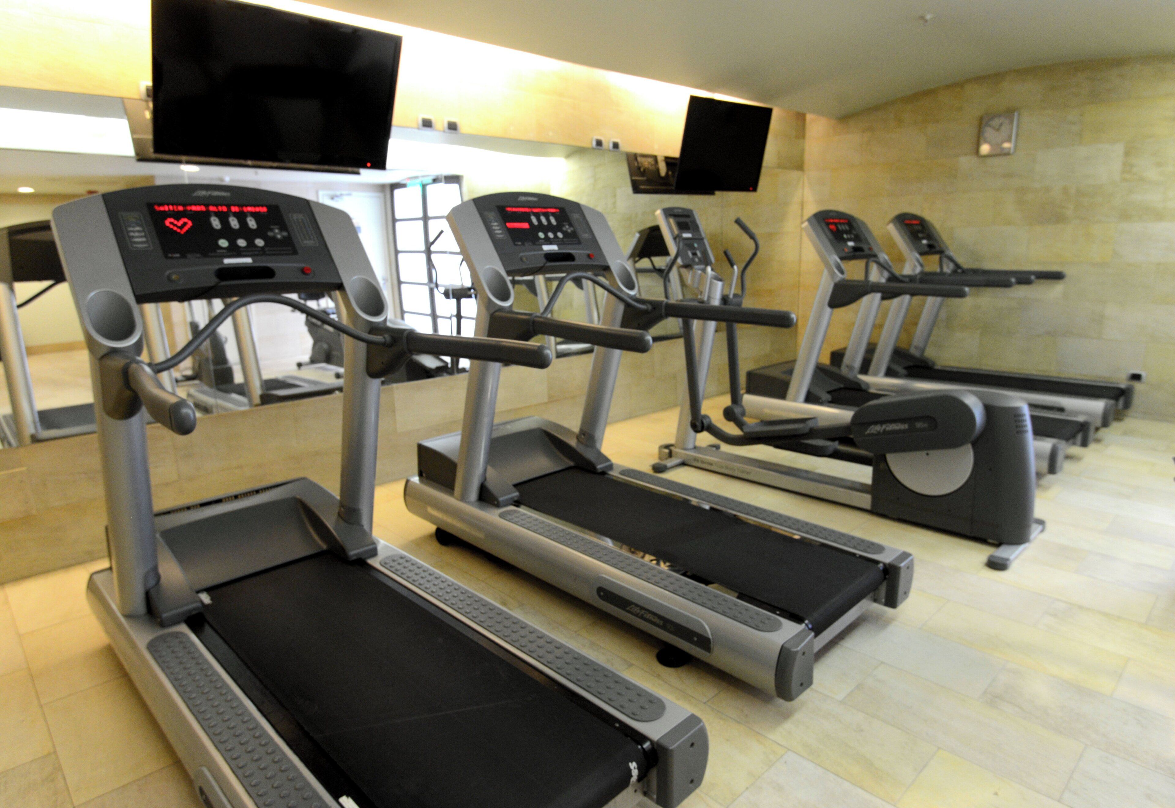 Varias máquinas de ejercicio estáticas en un gimnasio con piso de baldosas y un reloj en la pared.