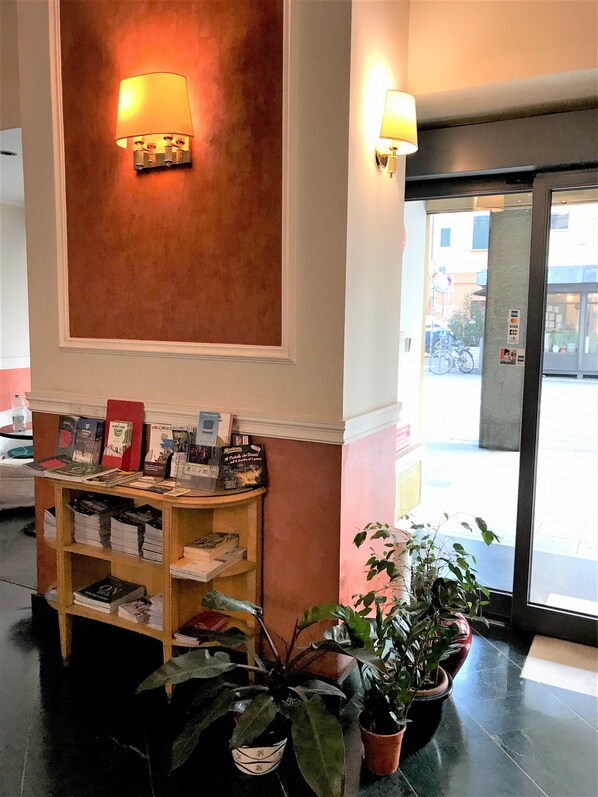 Una zona de recepción con un escritorio de madera que muestra libros y folletos, una lámpara de pared y plantas en macetas.