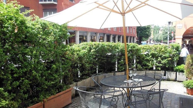 An outdoor patio with a round metal table and chairs, a large umbrella, and potted plants. - Hotel San Giorgio (Bologna)