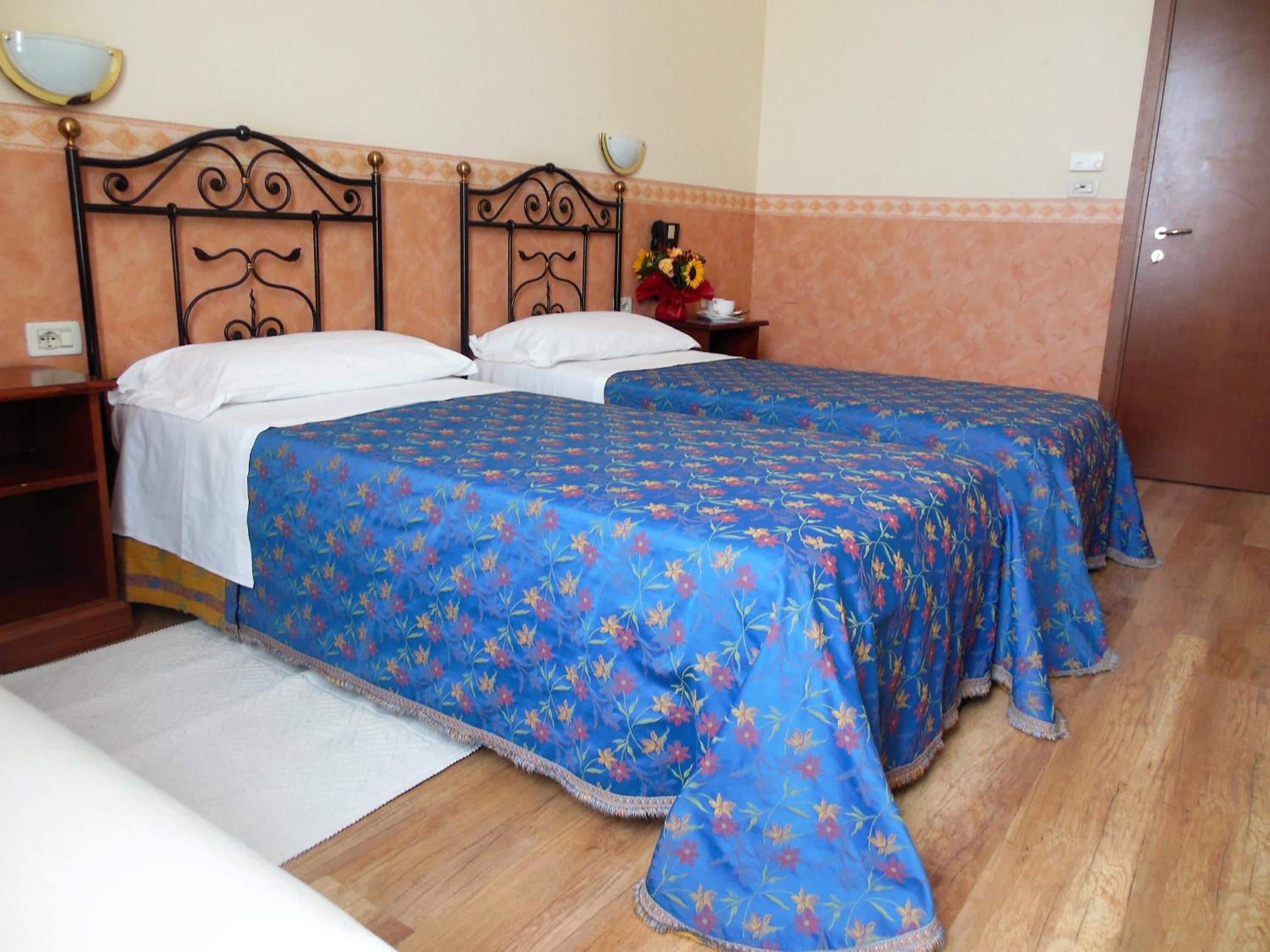 A room with two beds, each with a blue floral bedspread, a wooden headboard, and a nightstand with a vase of flowers.