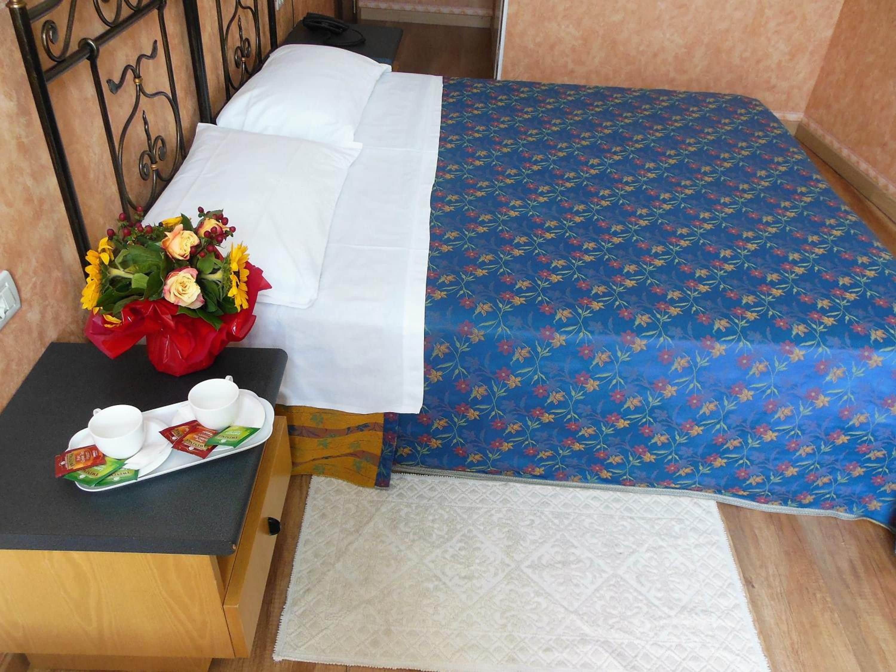 A neatly made bed with a blue floral bedspread, white pillows, and a bouquet of flowers on a bedside table.