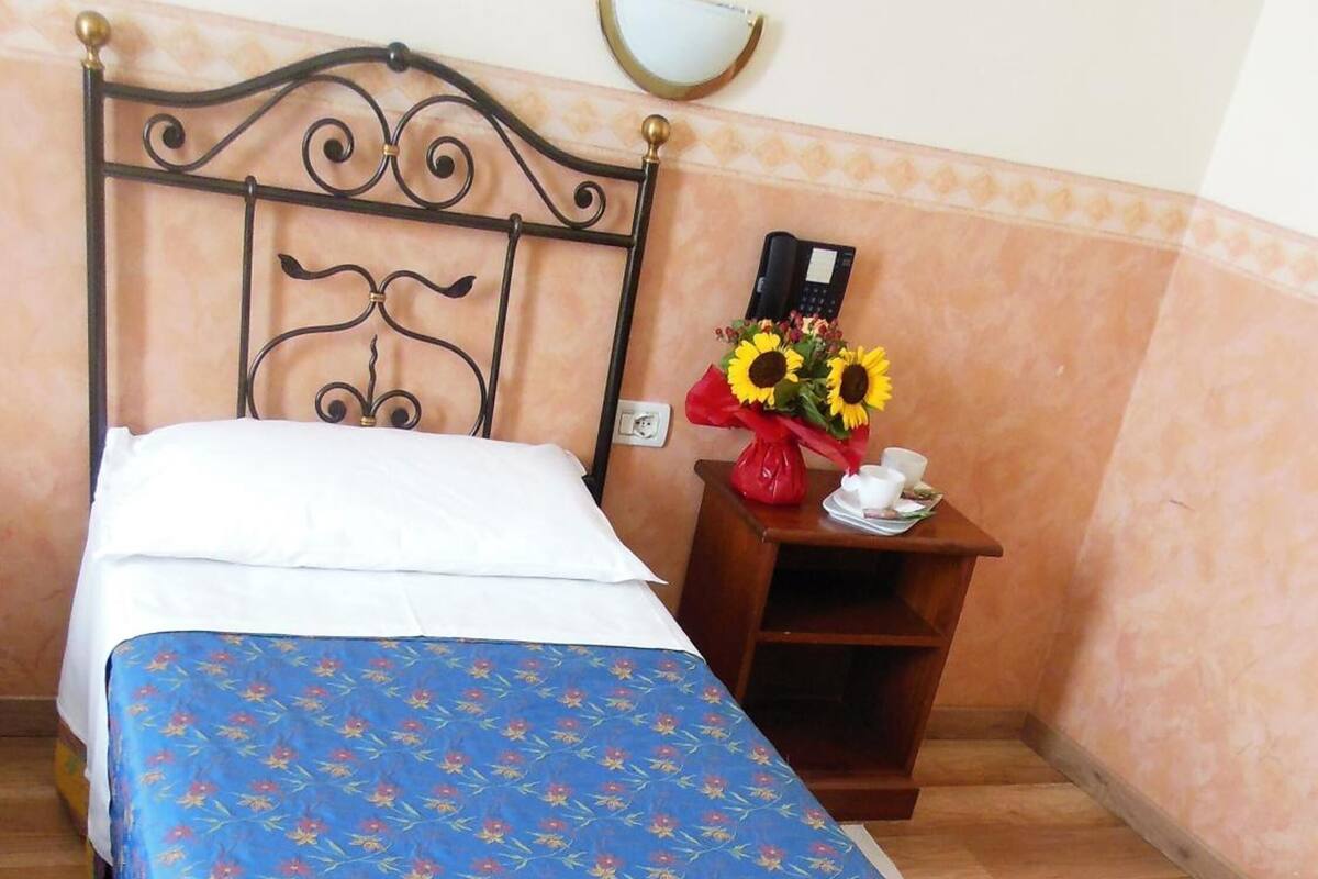 Ein Bett mit floralem Überwurf, ein Nachttisch mit Telefon und Blumenvase sowie eine an der Wand befestigte Lampe.