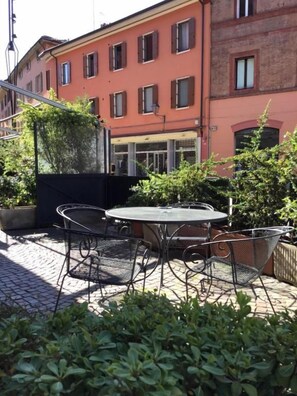 An outdoor patio with a round table and metal chairs, surrounded by greenery and a red building in the background. - Hotel San Giorgio (Bologna)