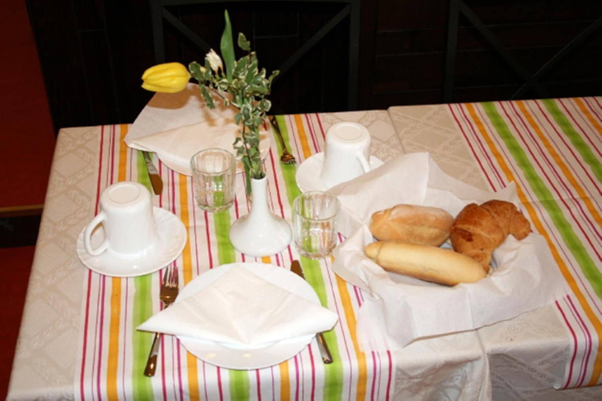 A table set with white plates, cups, and a small vase with flowers. There are two baguettes and a croissant on the table.