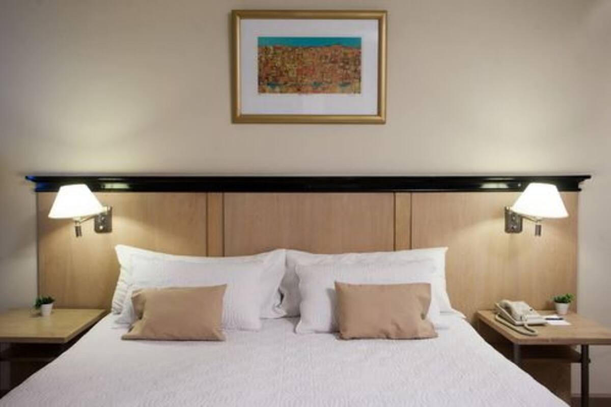 A neatly made bed with white and beige pillows, two bedside lamps, a framed artwork on the wall, and a wooden headboard.