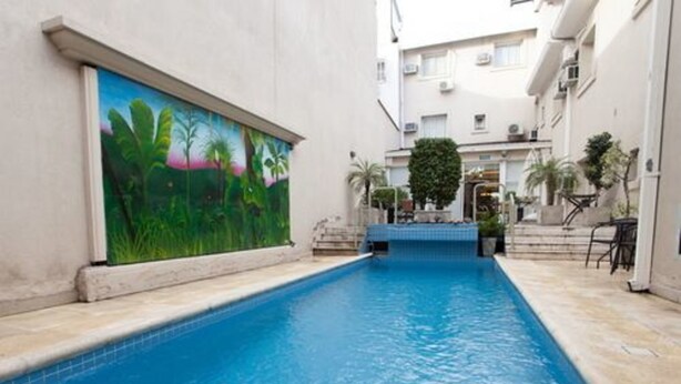 A swimming pool with a mural of tropical plants on one side, surrounded by a building with a tiled floor and a small garden area. - Argentino Hotel (Mendoza)