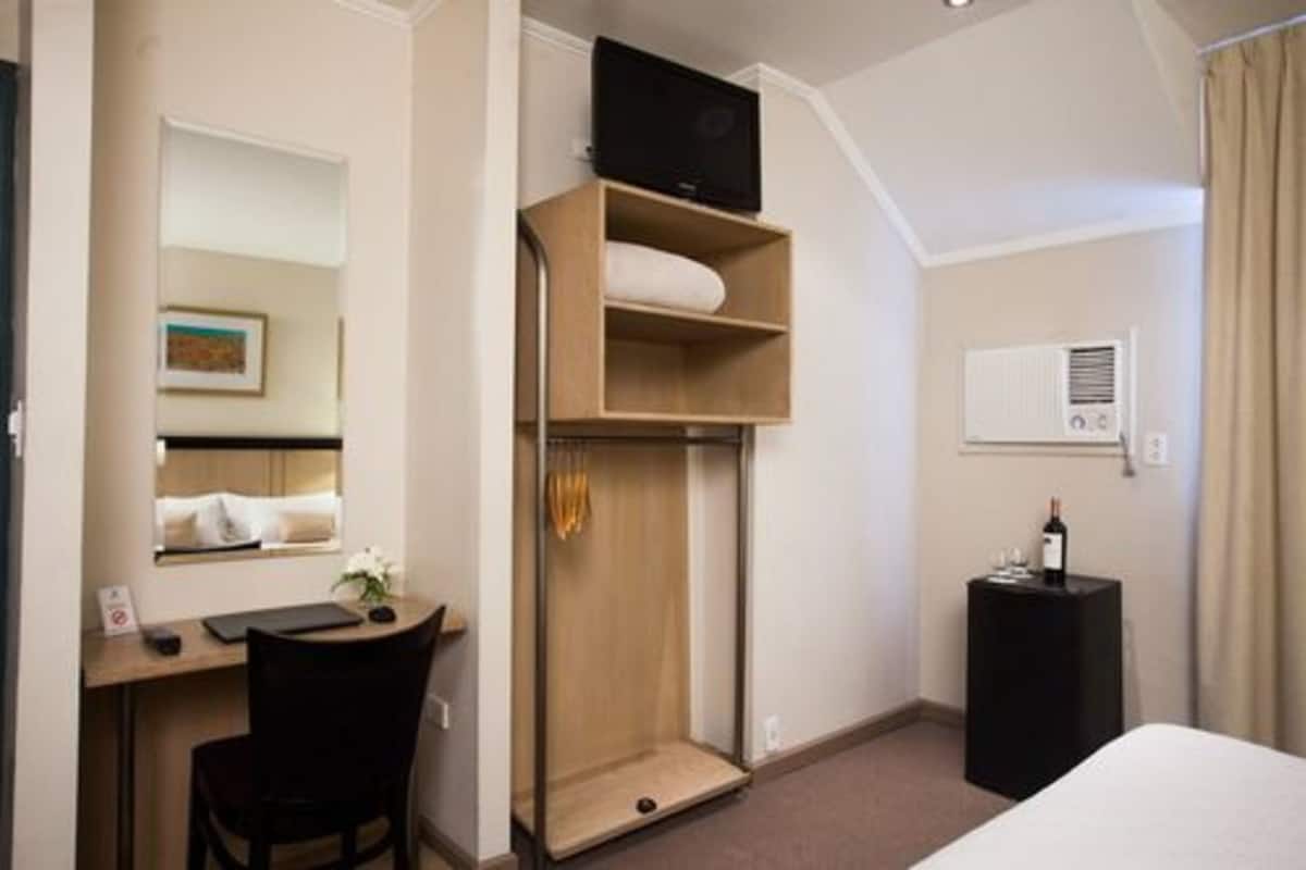 A hotel room with a bed, a desk with a chair, a wardrobe, a television on a shelf, a small table with a bottle, and a wall-mounted air conditioner.