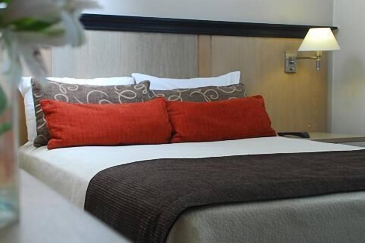 A hotel room with a bed, pillows, a nightstand, a lamp, and a vase with flowers.