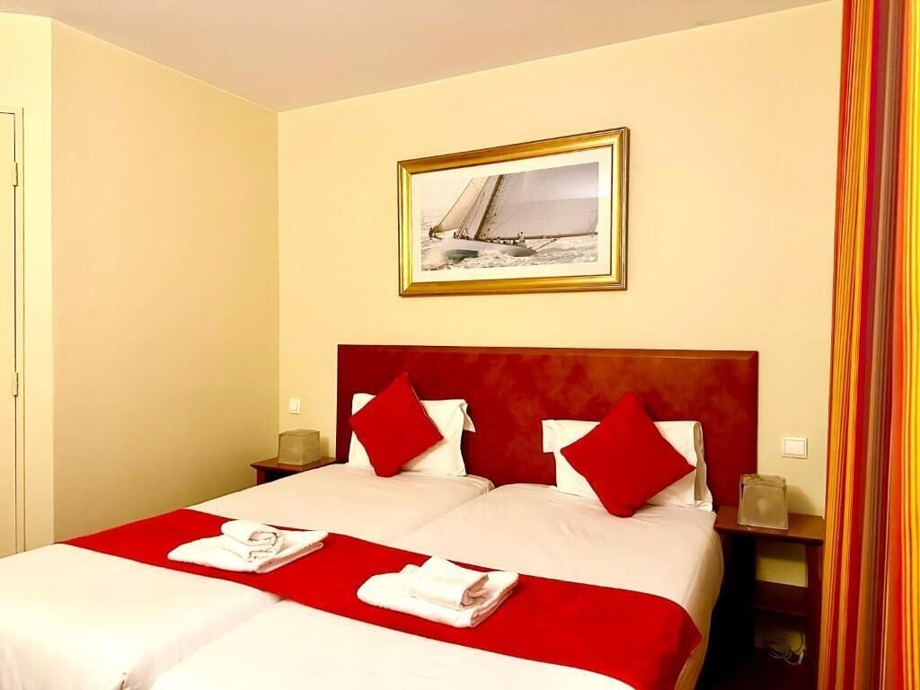 Habitación de hotel con una cama grande, ropa de cama roja, dos almohadas, cabecera, mesita de noche, un cuadro en la pared y una ventana con cortinas.