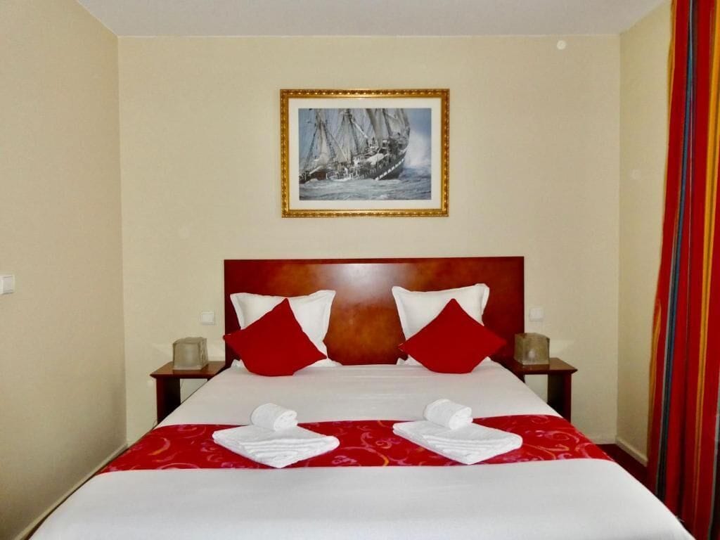 A hotel room with a large bed, red and white pillows, a framed picture on the wall, and a red curtain.
