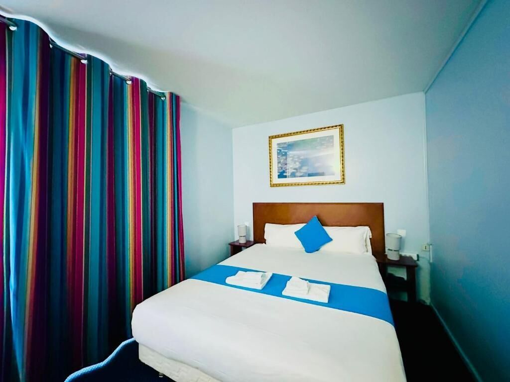 A hotel room with a large bed, a blue accent pillow, a framed picture on the wall, and a colorful curtain.