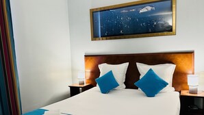 Een hotelkamer met een groot bed, twee kussens, een hoofdeinde, twee nachtkastjes met lampen, een aan de muur hangend schilderij en een raam met gordijnen.