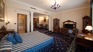 A hotel room with a large bed, a desk with a television, a chair, and a desk lamp. There is a door leading to a bathroom and a hallway. - Grand Hotel London (Varna)