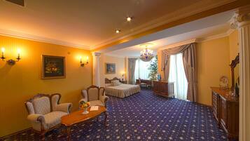 Une chambre d’hôtel avec un grand lit, deux fauteuils, une table en bois avec des fruits, un bureau et un lustre.