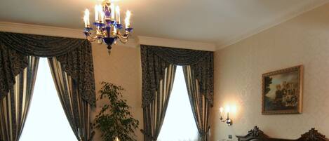 A hotel room with a bed, a desk, a chair, a lamp, a chandelier, and a painting on the wall.