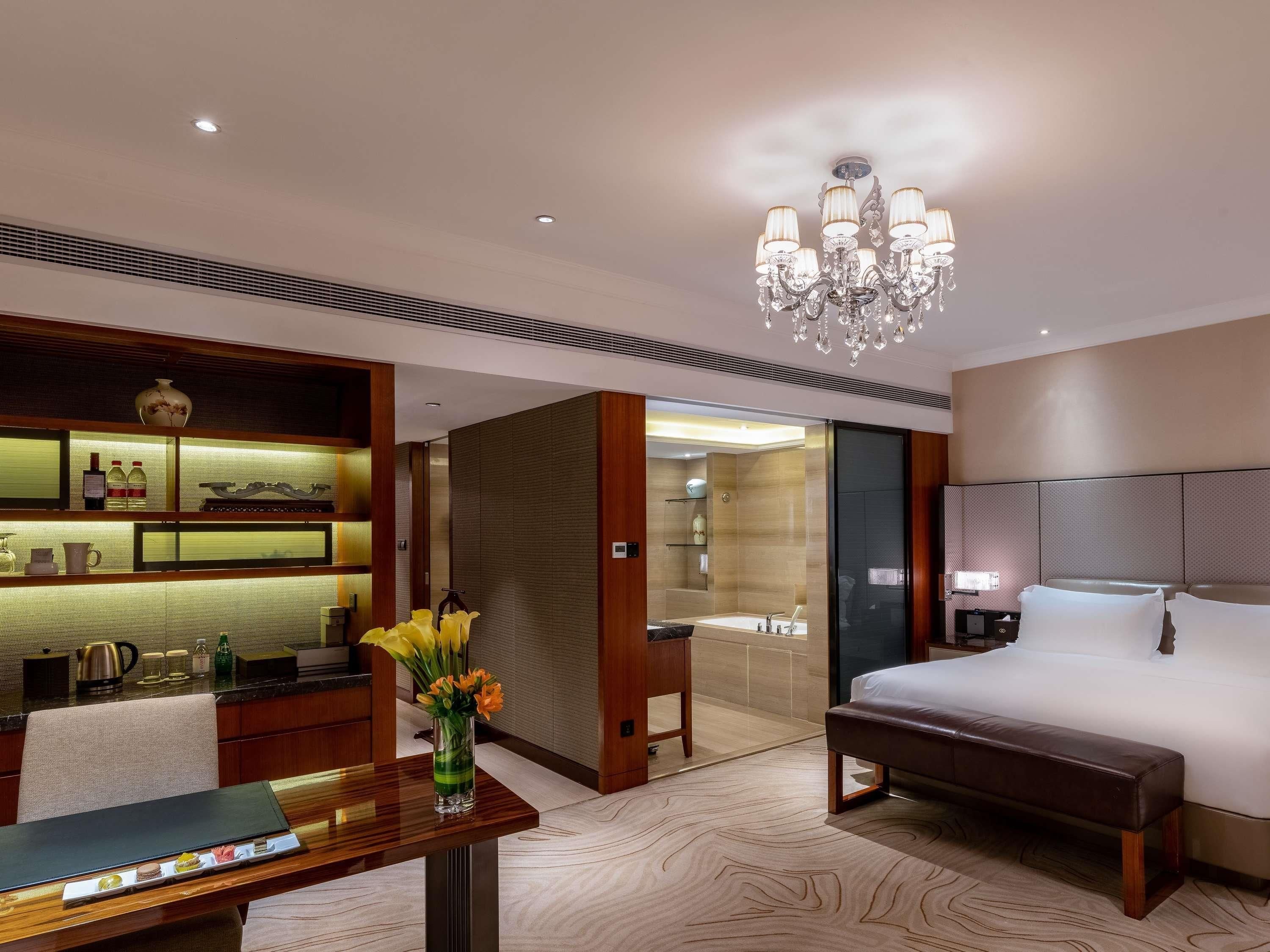 A hotel room with a bed, a desk, a vase with flowers, a chandelier, and a bathroom visible through an open door.