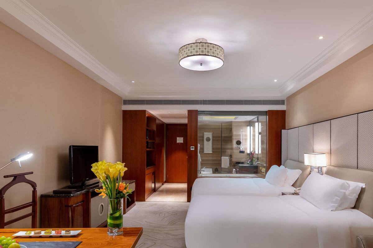 Ein Hotelzimmer mit einem Bett, einem Schreibtisch mit Lampe, einem Fernseher und einer Vase mit Blumen.