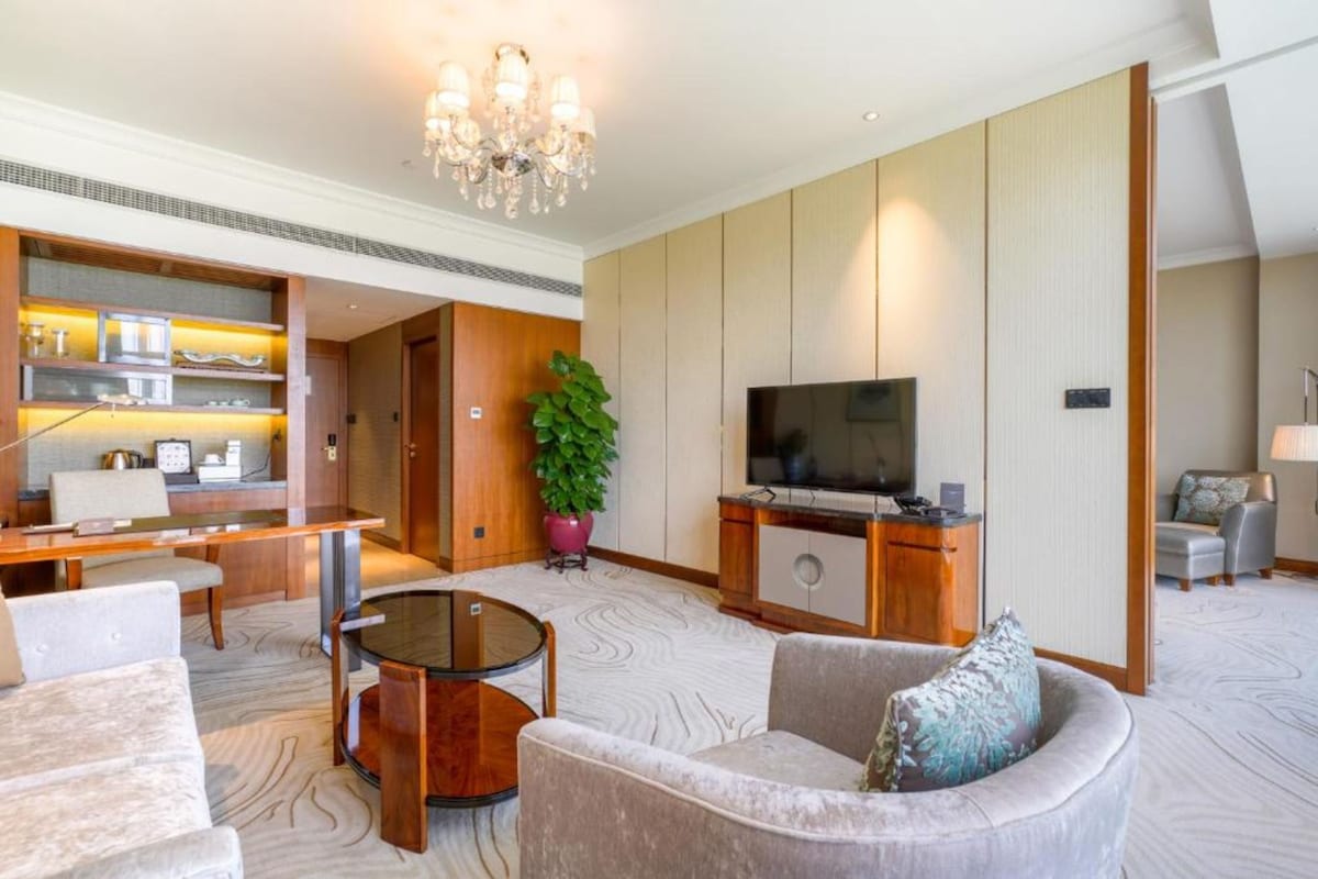 Een ruime woonkamer met een grote flatscreen-tv, comfortabele zitplaatsen, een kroonluchter en een potplant.