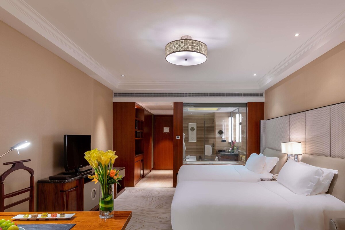 Une chambre d’hôtel avec un lit, un bureau avec une lampe, une télévision et un vase avec des fleurs.
