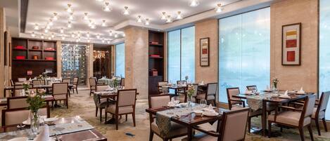 A spacious dining area with multiple tables and chairs, a chandelier, and large windows.