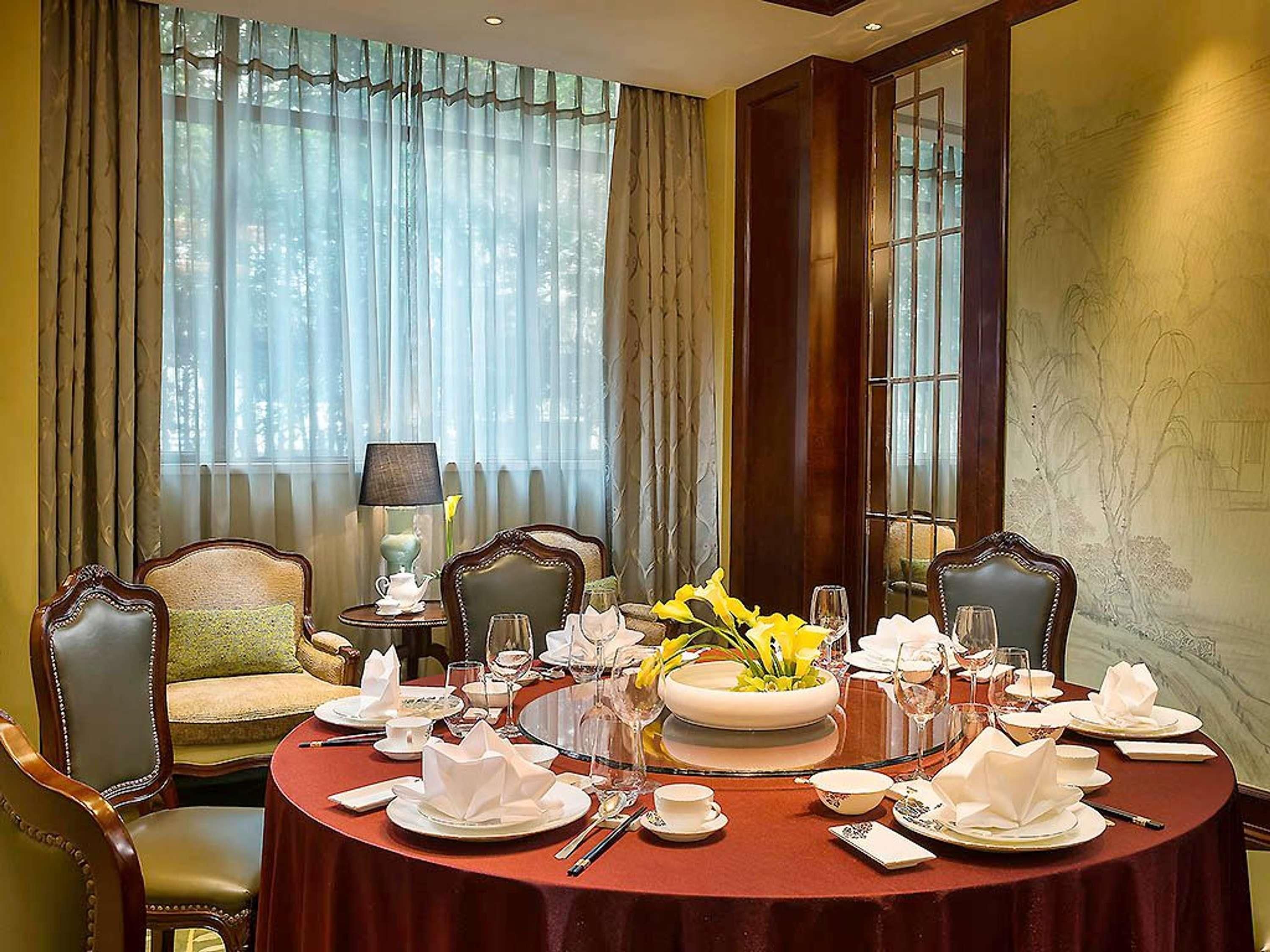 Uma sala de jantar formal com uma mesa redonda posta para seis pessoas, grandes janelas com cortinas e móveis clássicos.