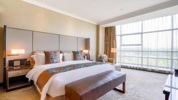 Ein Hotelzimmer mit einem großen Bett, einem Schreibtisch, einem Sessel und Blick ins Freie durch große Fenster.