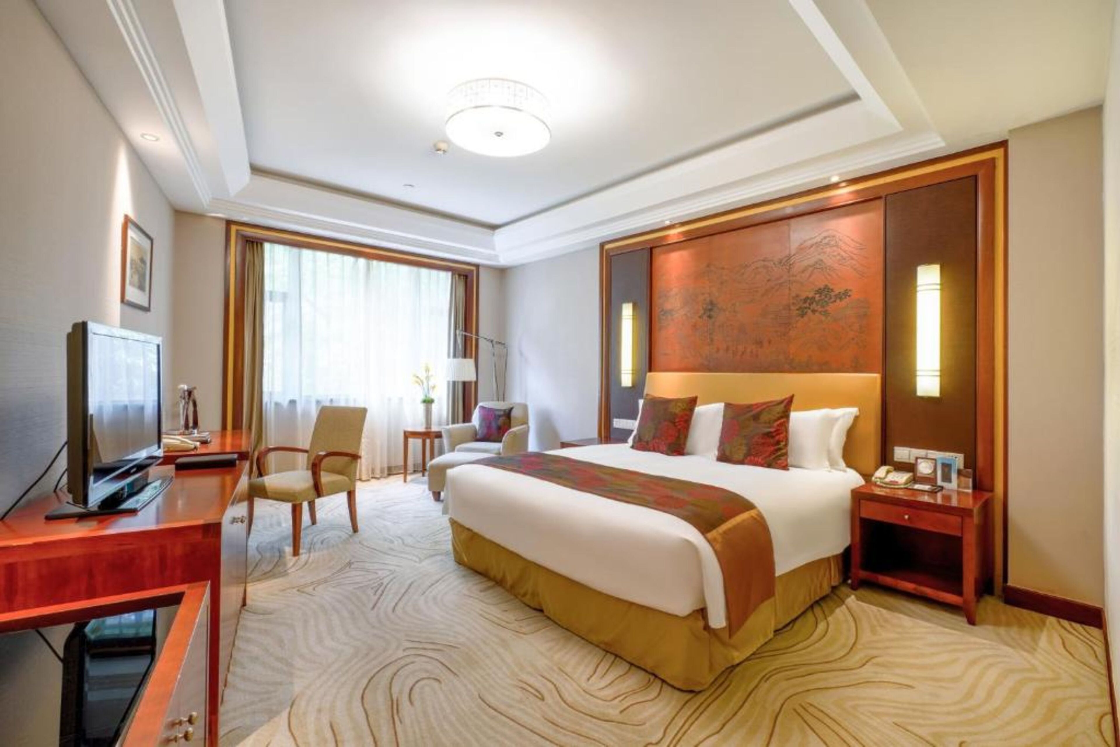 A hotel room with a large bed, a desk with a television, a chair, and a decorative wall panel.
