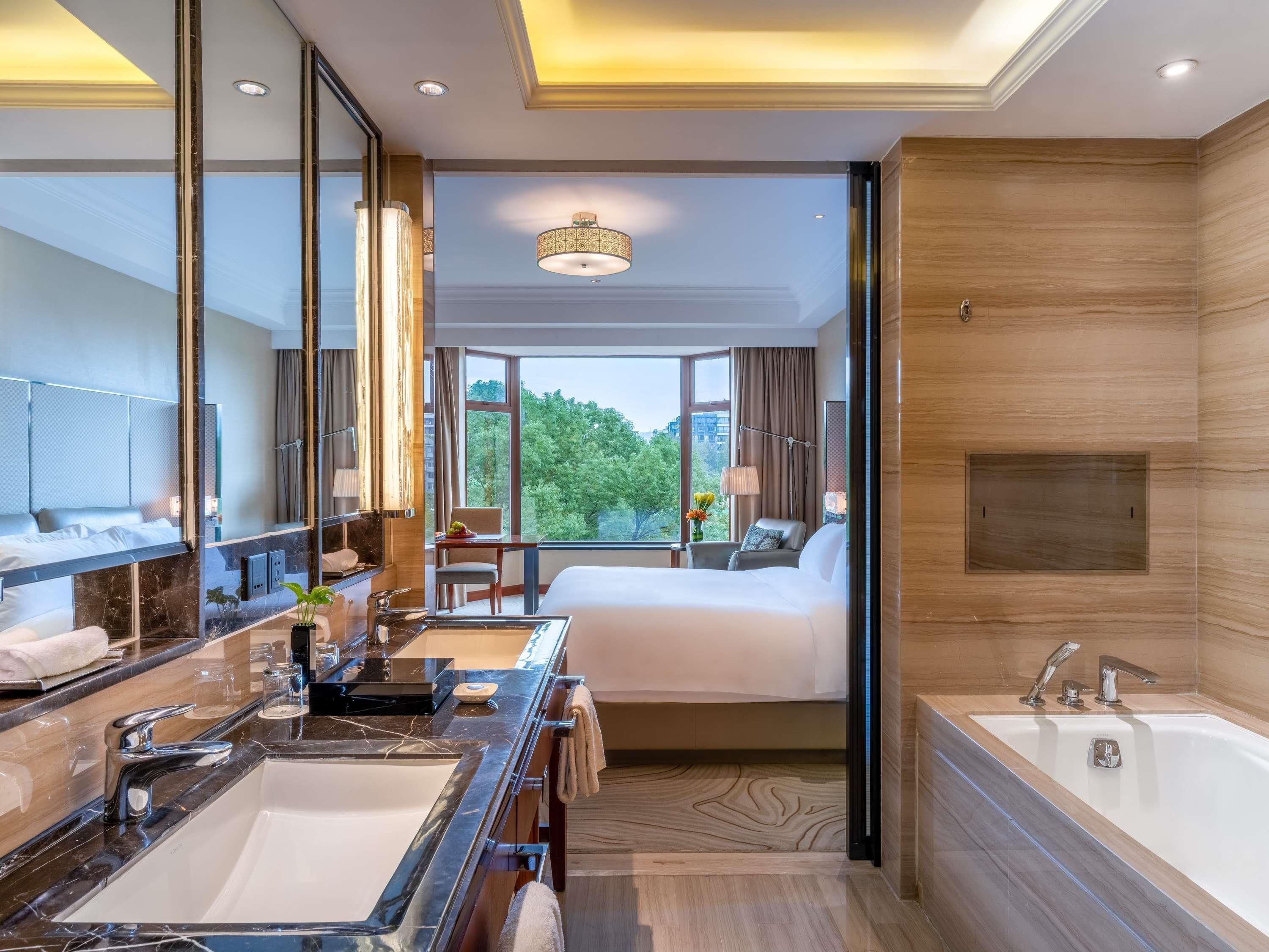Una habitación de hotel moderna con una cama grande, un baño independiente con bañera, tocador doble y un espejo amplio.
