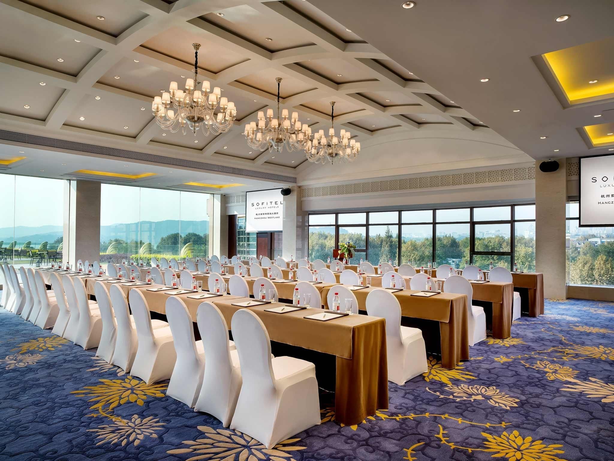 Un salón de conferencias con mesas y sillas, amplias ventanas, candelabros y suelo alfombrado.