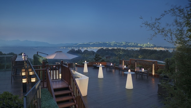 Una terraza de madera con mesas y sillas, con vistas panorámicas a montañas y una masa de agua.