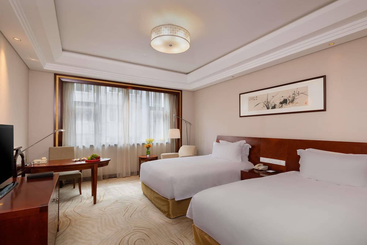 Ein Hotelzimmer mit zwei Betten, einem Schreibtisch, einem Fernseher, einer Lampe und einem Vorhangfenster.