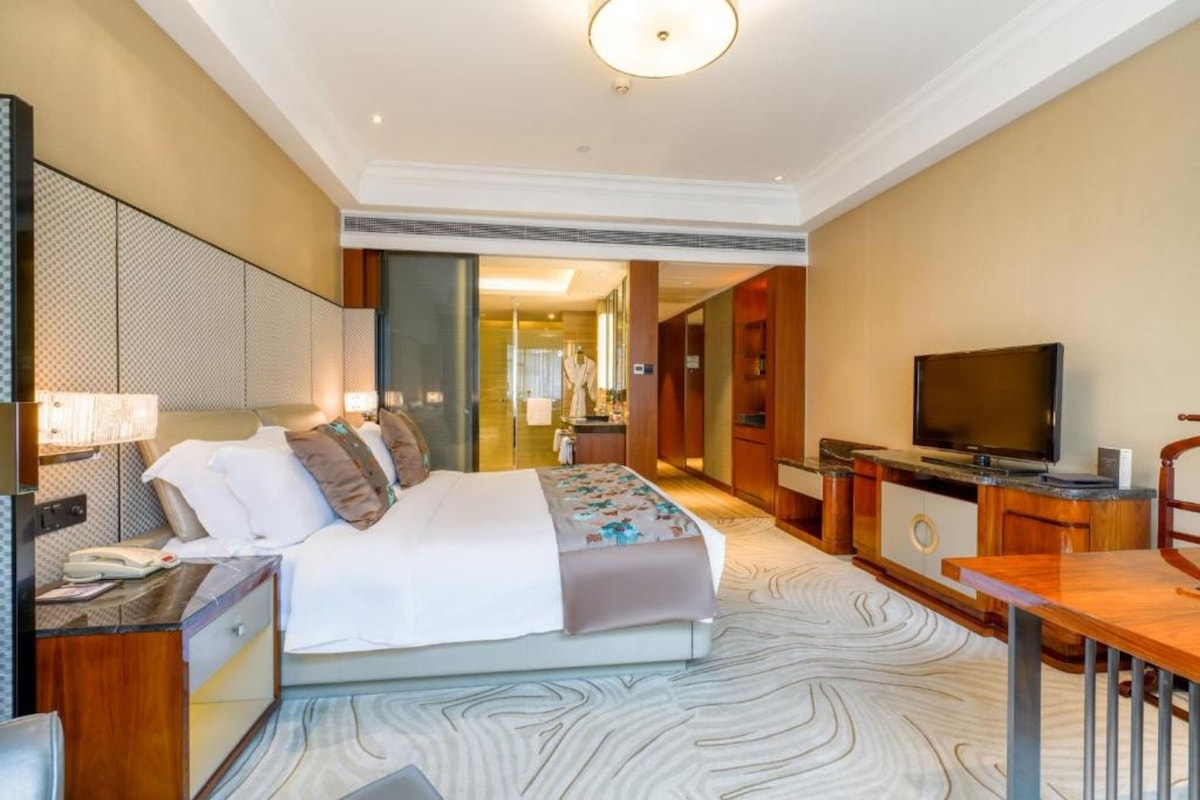 Een hotelkamer met een groot bed, een bureau met een lamp, een televisie en uitzicht op de gang.