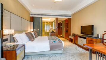 A hotel room with a large bed, a desk with a lamp, a television, and a view of the hallway.