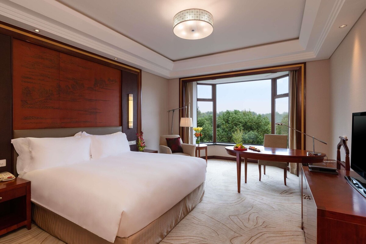 Une chambre d’hôtel avec un grand lit, un bureau, une chaise, une télévision et une fenêtre donnant sur des arbres.