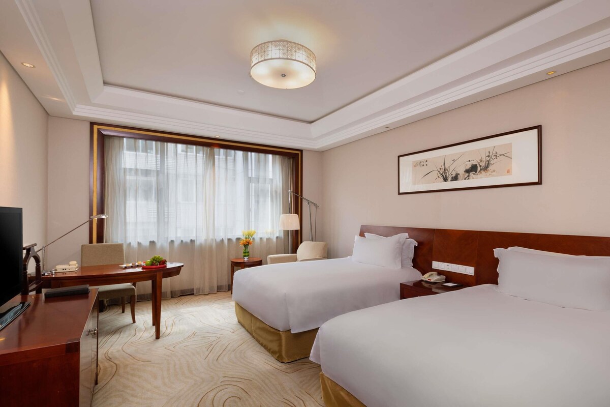 Une chambre d’hôtel avec deux lits, un bureau, une télévision, une lampe et une fenêtre avec des rideaux.
