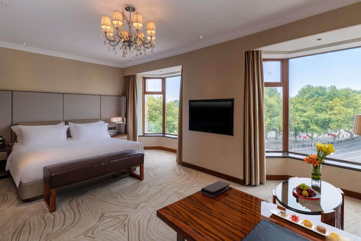 Ein Hotelzimmer mit einem großen Bett, einem an der Wand montierten Fernseher, einem Esstisch mit Obstschale und Blick auf Bäume durch das Fenster.