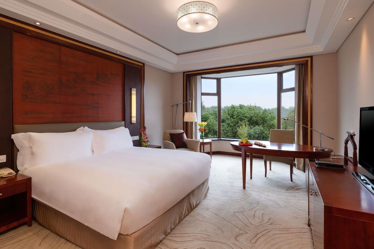 Ein Hotelzimmer mit einem großen Bett, einem Schreibtisch, einem Sessel, einem Fernseher und einem Fenster mit Blick auf Bäume.