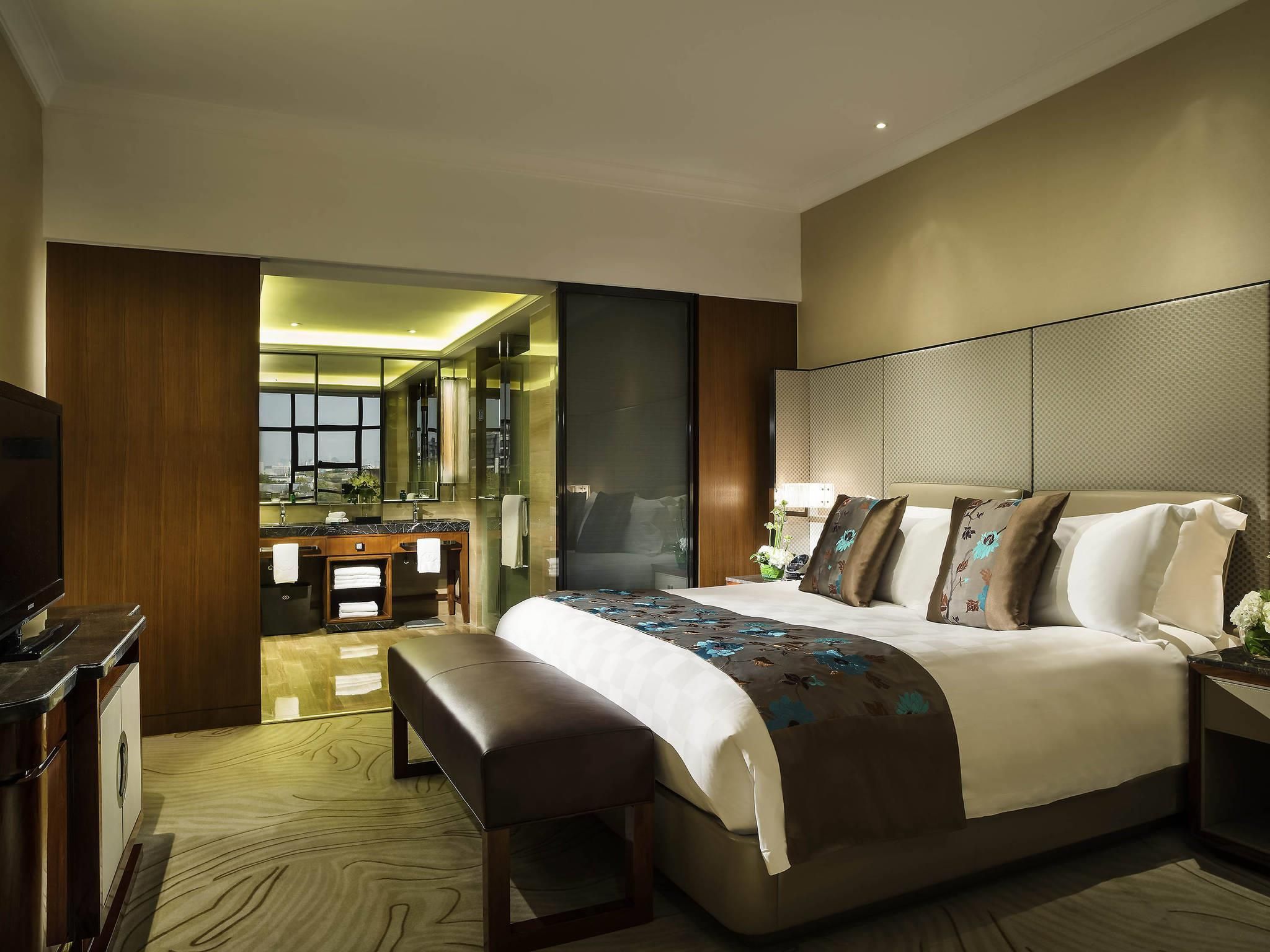 A modern hotel room with a large bed, a bench, a TV, a bathroom with a mirror, and a view of the city.