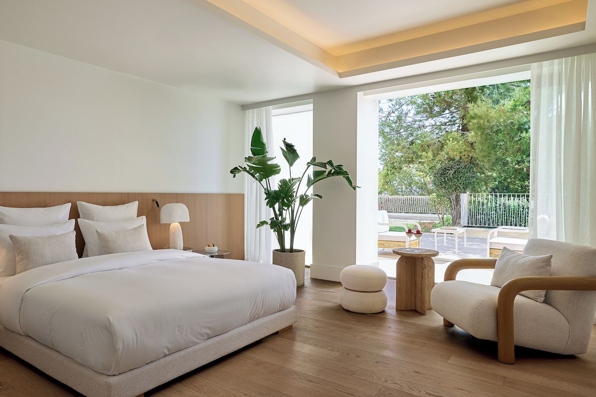 A modern hotel room with a large bed, a seating area with a wooden coffee table, and a view of the outdoors.