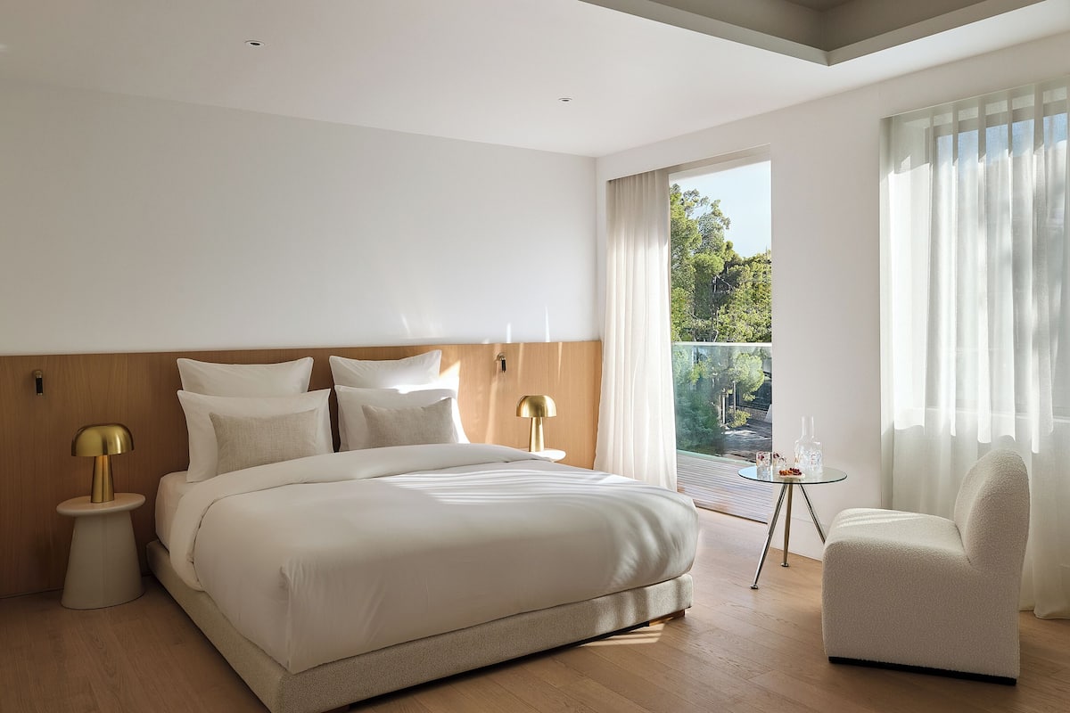 A modern hotel room with a large bed, two bedside tables with lamps, a chair, and a balcony with curtains.
