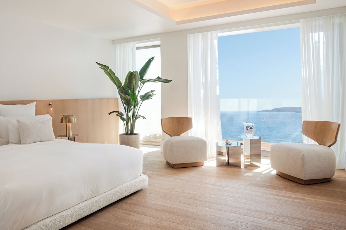 A modern hotel room with a large bed, two armchairs, a small table, and a potted plant.
