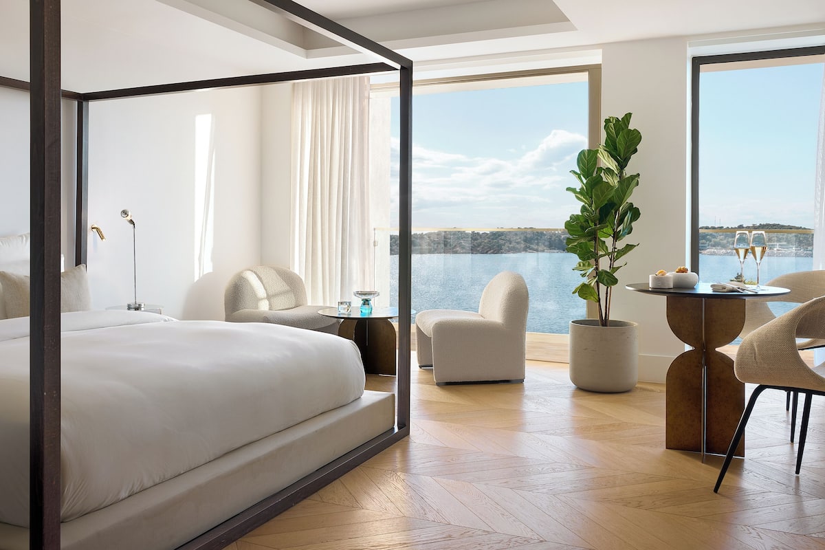 A modern hotel room with a large bed, a sitting area with a table, and a view of the sea.