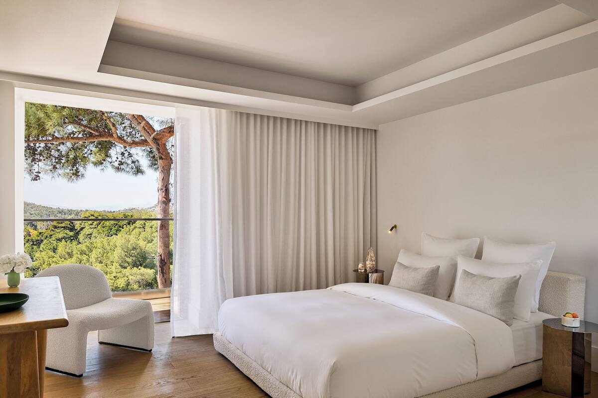 A bedroom with a large bed, a chair, a small table, and a view of greenery through a window.