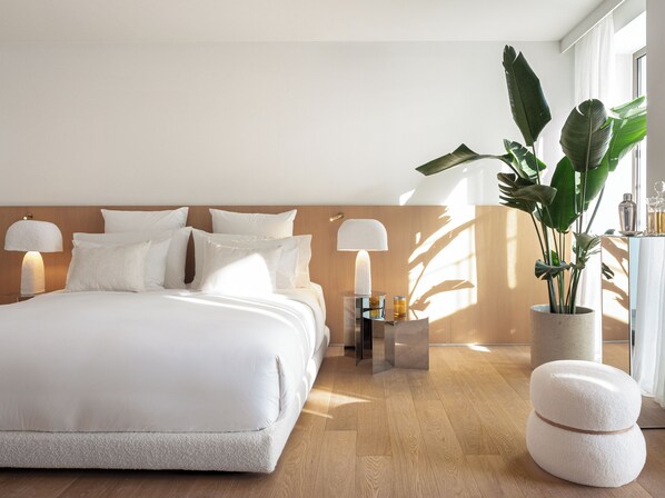 A modern bedroom with a large bed, bedside lamps, a nightstand, a potted plant, and a small table with a bottle and glass.