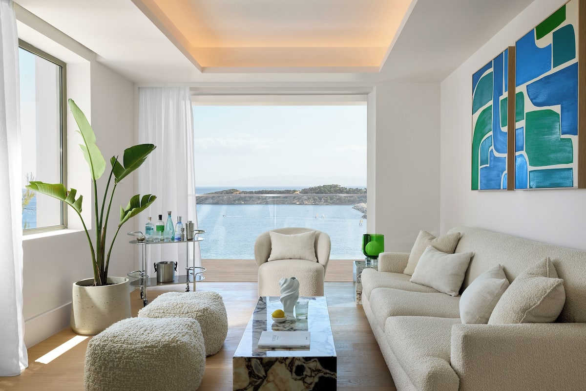 A modern living room with a large window, a sofa, ottomans, a coffee table, and a potted plant.