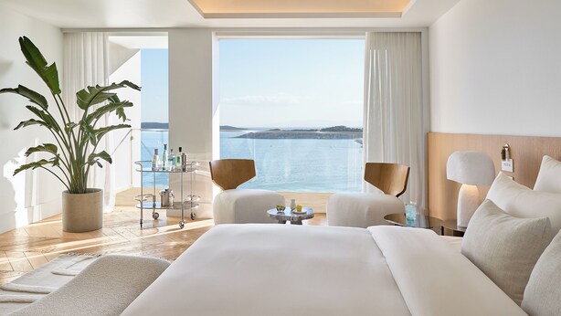 A modern hotel room with a large bed, two chairs, a small table, a potted plant, and a view of the sea.