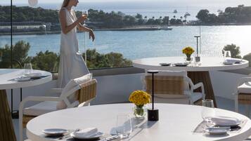 A dining area with tables set for two, overlooking a body of water with boats and a distant shoreline.
